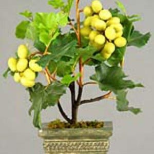 May include: A decorative artificial plant featuring a small tree with green leaves and clusters of yellow grapes. The plant is potted in a square, aged-green planter with decorative trim. The overall height is approximately 18 inches.