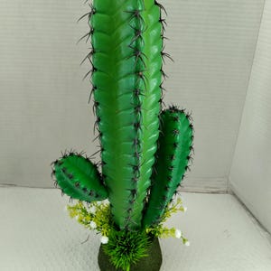 Op de afbeelding: Een felgroene kunstcactus met donkere stekels. De cactus heeft drie stengels en staat in een ronde, met mos bedekte basis met kleine witte en gele bloemen. Een decoratieve plant voor thuis of op kantoor.
