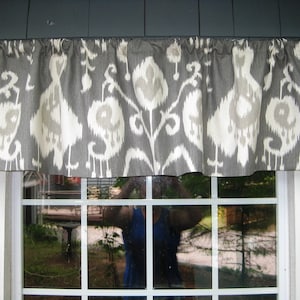 May include: A gray and white patterned valance is displayed above a window. The fabric features a repeating design of stylized floral shapes in light gray and white. The valance is gathered at the top and hangs in soft folds, adding a decorative touch to the window.