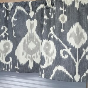 May include: Gray and cream-colored valance with an ikat pattern. The fabric features a repeating design of stylized floral and geometric shapes. The valance is gathered at the top and hangs over a window.