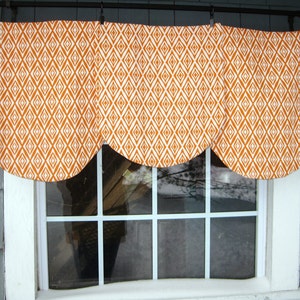 May include: A white window with a valance made of orange and white fabric with a diamond pattern. The valance is gathered in the center and has a scalloped edge.