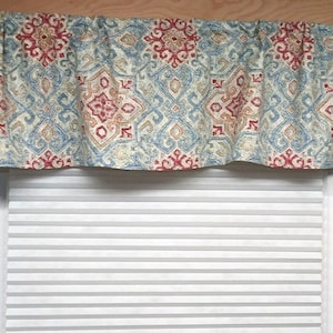 May include: A valance with a blue, red, and beige geometric pattern. The valance is made of fabric and has a ruffled edge. It is hanging over a white window blind.