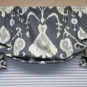 May include: A gray and cream-colored valance with an ikat pattern. The fabric is gathered at the top and draped at the sides, creating a soft, curved shape. The valance is designed to be mounted above a window.