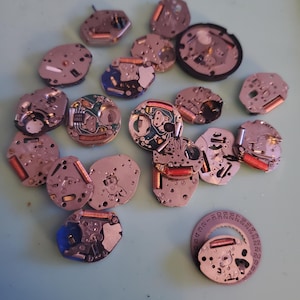 May include: A collection of disassembled watch movements, featuring various shapes and sizes. The components display a mix of silver, copper, and blue hues, with intricate internal mechanisms visible. Some pieces have red and copper-colored coils.