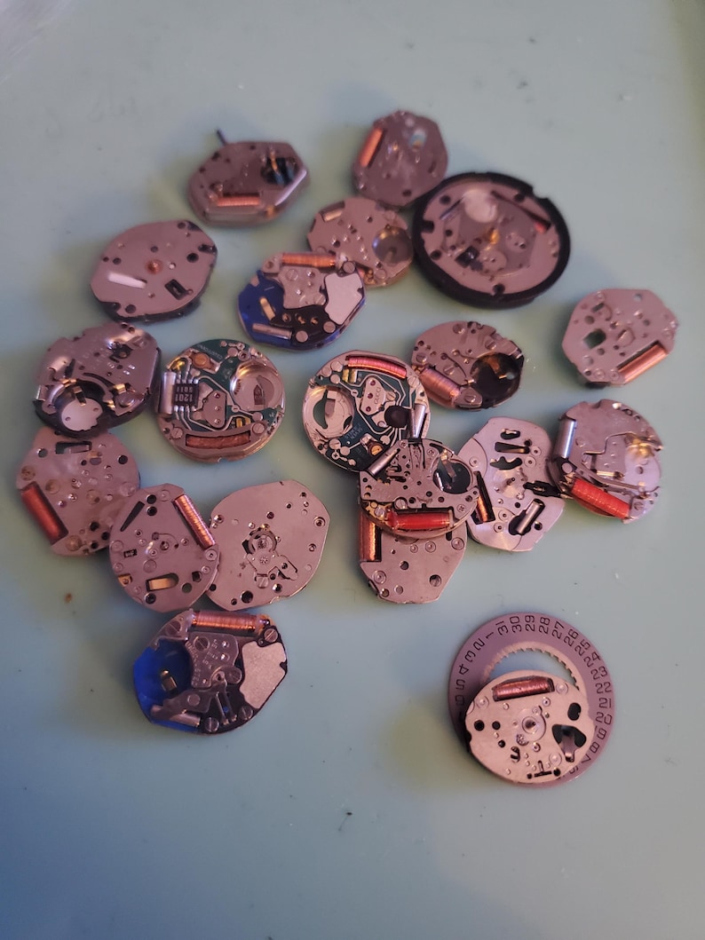 May include: A collection of disassembled watch movements, featuring various shapes and sizes. The components display a mix of silver, copper, and blue hues, with intricate internal mechanisms visible. Some pieces have red and copper-colored coils.