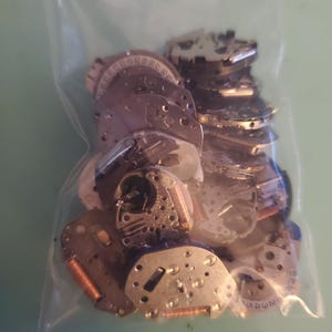 May include: A clear plastic bag filled with various small, silver-colored watch parts. The parts include circular and rectangular components with intricate details, gears, and numbers. Some parts have copper-colored coils. The bag is on a light green surface.
