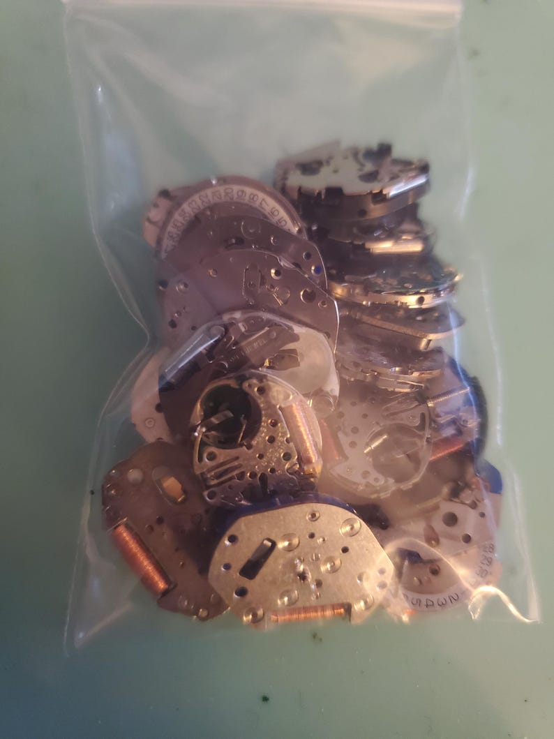 May include: A clear plastic bag filled with various small, silver-colored watch parts. The parts include circular and rectangular components with intricate details, gears, and numbers. Some parts have copper-colored coils. The bag is on a light green surface.