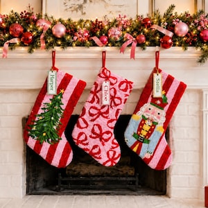 May include: Three textured Christmas stockings hanging from a mantel. The left stocking features a green tree and a tag labeled Harper. The center stocking has a pink bow pattern and a tag labeled Chloe. The right stocking shows a nutcracker and a tag labeled Stella.