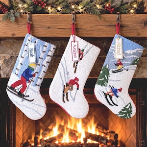 May include: Three needlepoint Christmas stockings hanging from a wooden mantel. Each stocking features a winter skiing scene with figures on snow and includes a rectangular wooden name tag labeled Noah, Avalane, or Michael.