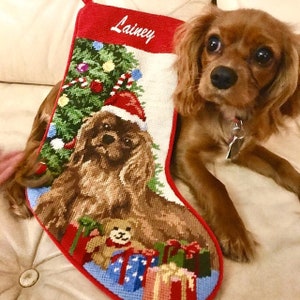 May include: A red and white Christmas stocking with a personalized name "Lainey" embroidered on the top. The stocking features a green Christmas tree with ornaments and candy canes, a brown dog wearing a Santa hat, and a teddy bear with presents.