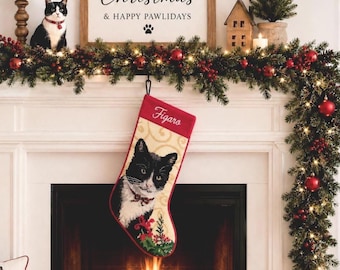 Puede incluir: Un calcetín navideño con una imagen tejida de un gato tuxedo blanco y negro con el nombre Figaro bordado en letra cursiva blanca en la parte superior. El calcetín tiene un fondo crema con un patrón de remolinos sutil y un diseño de acebo.