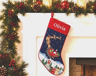 May include: A Christmas stocking featuring a dark blue body with a needlepoint design of Santa Claus in a sleigh pulled by reindeer flying over a snowy village. The top cuff is red with the name Olivia embroidered in white capital letters.