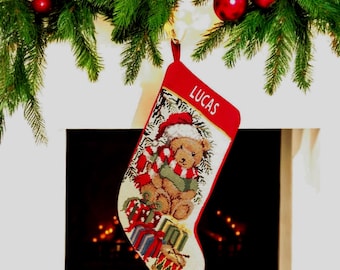 May include: A red Christmas stocking featuring a needlepoint design of a teddy bear wearing a Santa hat and scarf, surrounded by wrapped gifts. The name LUCAS is embroidered in white capital letters on the red cuff at the top of the stocking.
