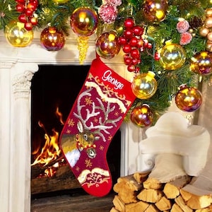 May include: Red Christmas stocking with a reindeer design and the name "George" embroidered on it. The stocking is hanging on a fireplace mantel decorated with ornaments and greenery.