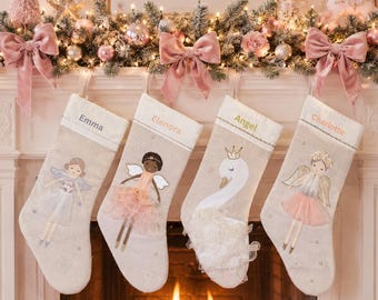 May include: Four beige fabric Christmas stockings hanging in a row. Each stocking features a personalized name at the top: Emma, Elenora, Angel, and Charlotte. The designs include embroidered fairy figures with tulle skirts and a swan with a crown.