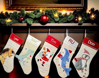 May include: Five beige Christmas stockings hanging in a row, each featuring a unique embroidered character: a horse, a dinosaur, Santa Claus, a shark, and a penguin. Each stocking has a red cuff with a name embroidered in white or gold thread.