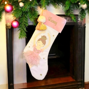 May include: A white Christmas stocking with a pink top and a ballerina design. The stocking features the name "Lainey" embroidered in gold. Gold stars and a pink tutu accent the ballerina.
