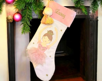 May include: A white Christmas stocking featuring a pink velvet cuff embroidered with the name Lainey. The stocking is decorated with a ballerina illustration wearing a pink tulle tutu and a gold crown, accented by small gold embroidered stars.