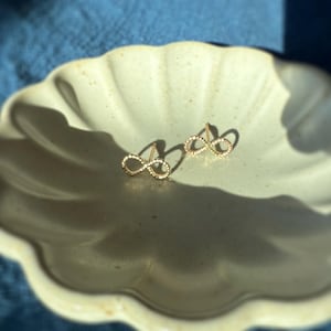 May include: A pair of gold-colored infinity symbol stud earrings displayed on a light-colored, scalloped ceramic dish. The earrings are small and delicate, with a subtle sparkle. The dish sits on a blue fabric surface, with sunlight casting shadows.