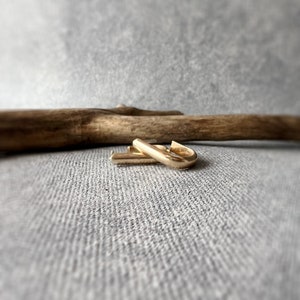 May include: A pair of gold-colored, rectangular hoop earrings. The earrings are displayed on a gray textured surface, with a piece of driftwood in the background. The earrings have a polished finish and a simple, modern design.