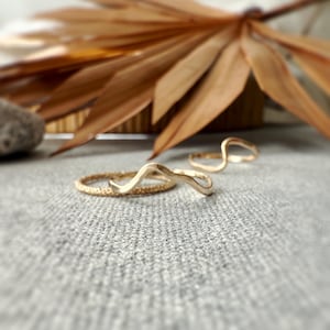 May include: Three gold rings on a gray textured surface. Two rings have a wavy design, and one is a textured band. A dried brown palm leaf is in the background, with a blurred focus.