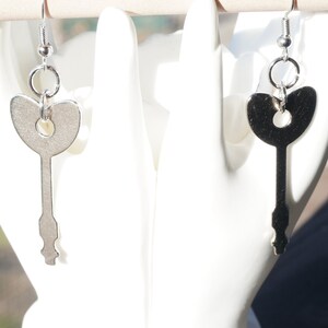 May include: Silver-toned key-shaped earrings. Each earring features a key design with a heart-shaped top and a detailed bottom. The earrings are suspended from a simple hook and ring closure. The background is a white surface.