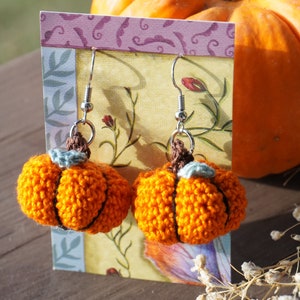 May include: A pair of crocheted pumpkin earrings with silver hooks. The earrings are orange and have a brown stem with a small green leaf.