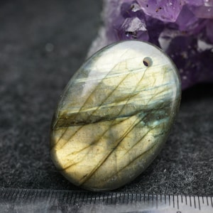 May include: A smooth, oval-shaped labradorite gemstone pendant with a hole drilled through the top for stringing. The stone has a greenish-blue hue with iridescent streaks of gold and silver.