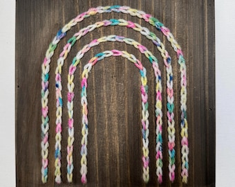 May include: A rainbow made of yarn is strung on a wooden board. The rainbow is made of five different colors: pink, blue, yellow, white, and green.