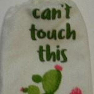 Peut inclure: Serviette de cuisine blanche avec un dessous de plat en crochet sur le dessus. La serviette est brodée de cactus verts et du texte "can't touch this".