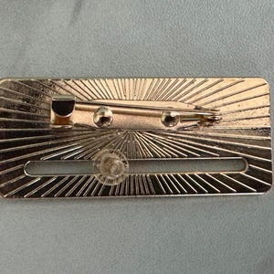 May include: A rectangular, gold-colored metal pin with a sunburst design. The pin features a safety clasp and a clear plastic fastener. The back of the pin is visible, showing the pin mechanism and the sunburst pattern.