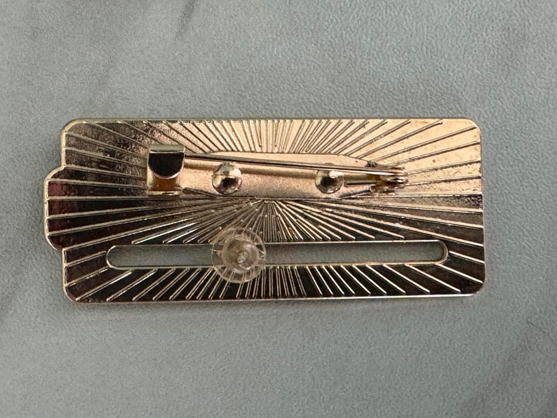 May include: A rectangular, gold-colored metal pin with a sunburst design. The pin features a safety clasp and a clear plastic fastener. The back of the pin is visible, showing the pin mechanism and the sunburst pattern.