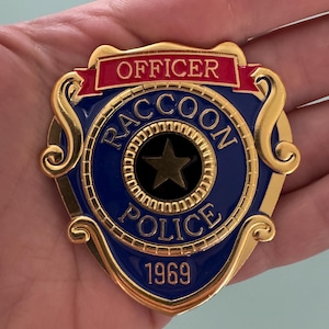 May include: A gold-colored metal badge with a blue center, featuring the words "OFFICER" and "RACCOON POLICE 1969." The badge has a star in the center and a decorative border.