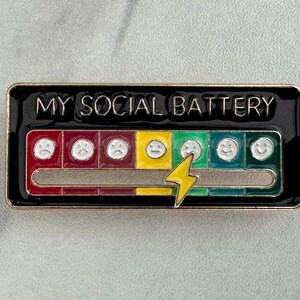 May include: A rectangular enamel pin with the text "MY SOCIAL BATTERY". The pin features a battery level indicator with a series of faces ranging from sad to happy, and a yellow lightning bolt. The pin has a black border.