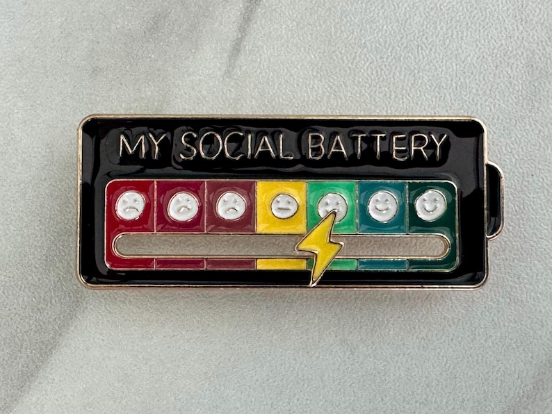 May include: A rectangular enamel pin with the text "MY SOCIAL BATTERY". The pin features a battery level indicator with a series of faces ranging from sad to happy, and a yellow lightning bolt. The pin has a black border.