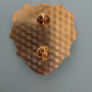 May include: Close-up of the back of a gold-colored pin with two pin backs. The pin has a textured, geometric pattern and a decorative edge. The pin is on a light blue background.