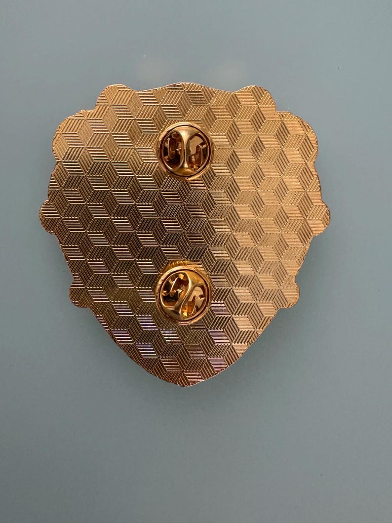 May include: Close-up of the back of a gold-colored pin with two pin backs. The pin has a textured, geometric pattern and a decorative edge. The pin is on a light blue background.