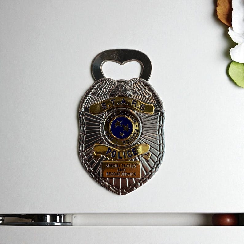 May include: Silver-colored bottle opener in the shape of a police badge. The badge features the text "S.T.A.R.S. Raccoon Police" and "Special Tactics and Rescue Service". The badge has a blue and gold color scheme.