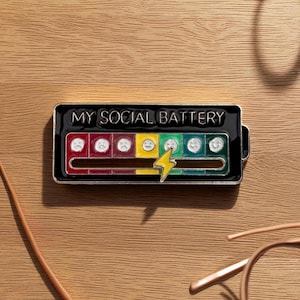 May include: A black enamel pin with the text "MY SOCIAL BATTERY" and a battery level indicator. The indicator features a series of emoji faces ranging from sad to happy, with a yellow lightning bolt. The pin is rectangular with a silver border.