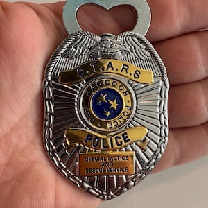 May include: Silver bottle opener in the shape of a police badge. The badge has gold accents and the text "S.T.A.R.S. Raccoon Police" and "Special Tactics and Rescue Service". The opener has a heart-shaped cutout.