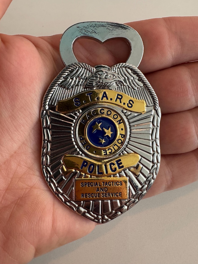 May include: Silver bottle opener in the shape of a police badge. The badge has gold accents and the text "S.T.A.R.S. Raccoon Police" and "Special Tactics and Rescue Service". The opener has a heart-shaped cutout.