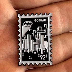 May include: A black and white Gotham city pin shaped like a postage stamp. The pin features the Batman logo and the word "Gotham". The pin has a white border and the price "92¢".