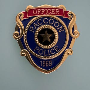 May include: A gold-colored metal badge with a blue center and red banner. The badge reads "OFFICER RACCOON POLICE 1969" with a gold star in the center. The badge has a decorative gold border.