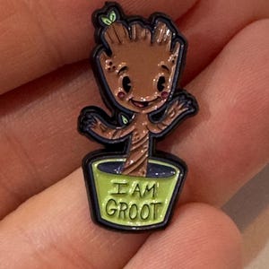 May include: A pin featuring a cartoon-style Groot character from Guardians of the Galaxy. The pin is brown with a black outline and has the words "I AM GROOT" in green on a green pot.