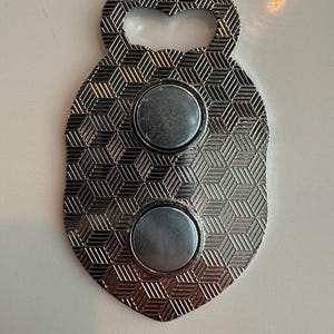 May include: Silver bottle opener with a shield shape and a geometric pattern. It has two circular magnets and a keyhole-shaped opening. The opener is made of metal and has a shiny finish.