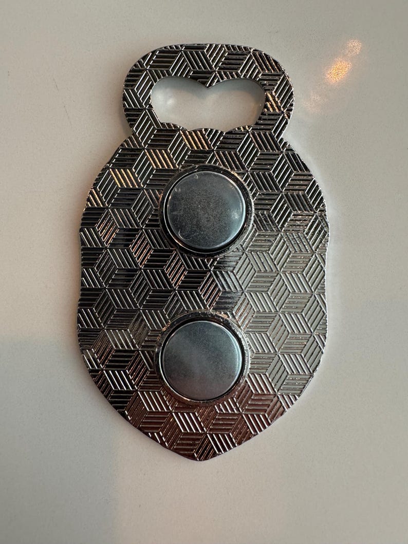 May include: Silver bottle opener with a shield shape and a geometric pattern. It has two circular magnets and a keyhole-shaped opening. The opener is made of metal and has a shiny finish.