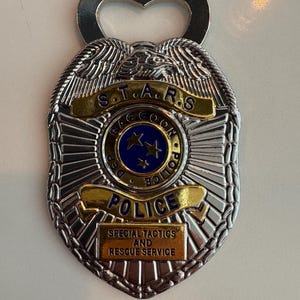 May include: Silver-colored bottle opener in the shape of a police badge. The badge has the text "S.T.A.R.S. RACCOON POLICE DEPT" and "SPECIAL TACTICS AND RESCUE SERVICE". The center of the badge has a blue circle with three stars.
