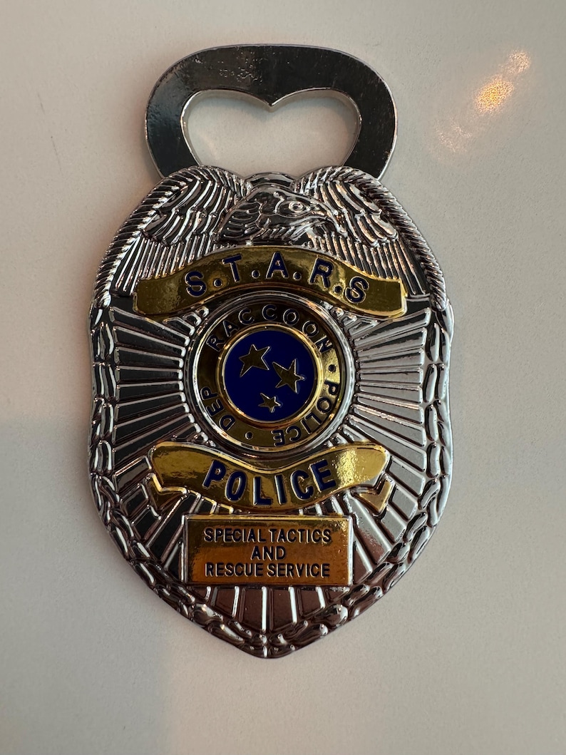 May include: Silver-colored bottle opener in the shape of a police badge. The badge has the text "S.T.A.R.S. RACCOON POLICE DEPT" and "SPECIAL TACTICS AND RESCUE SERVICE". The center of the badge has a blue circle with three stars.