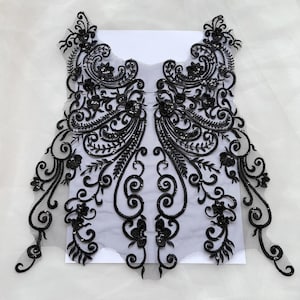 May include: A black lace applique featuring intricate floral and scrollwork embroidery with small black sequins. The design is symmetrical, crafted on a sheer black mesh base, and displays detailed swirling patterns and leaf-like motifs throughout.