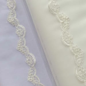 May include: Two strips of white lace trim featuring a scalloped edge with a repeating floral and leaf pattern. The lace is layered over sheer white fabric, showcasing the intricate embroidery and delicate threadwork of the floral design.